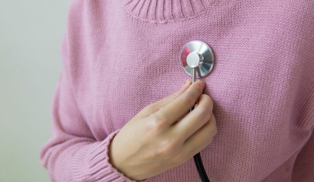 A person wearing a pink sweater holds a stethoscope to their chest, suggesting a health check or self-examination.